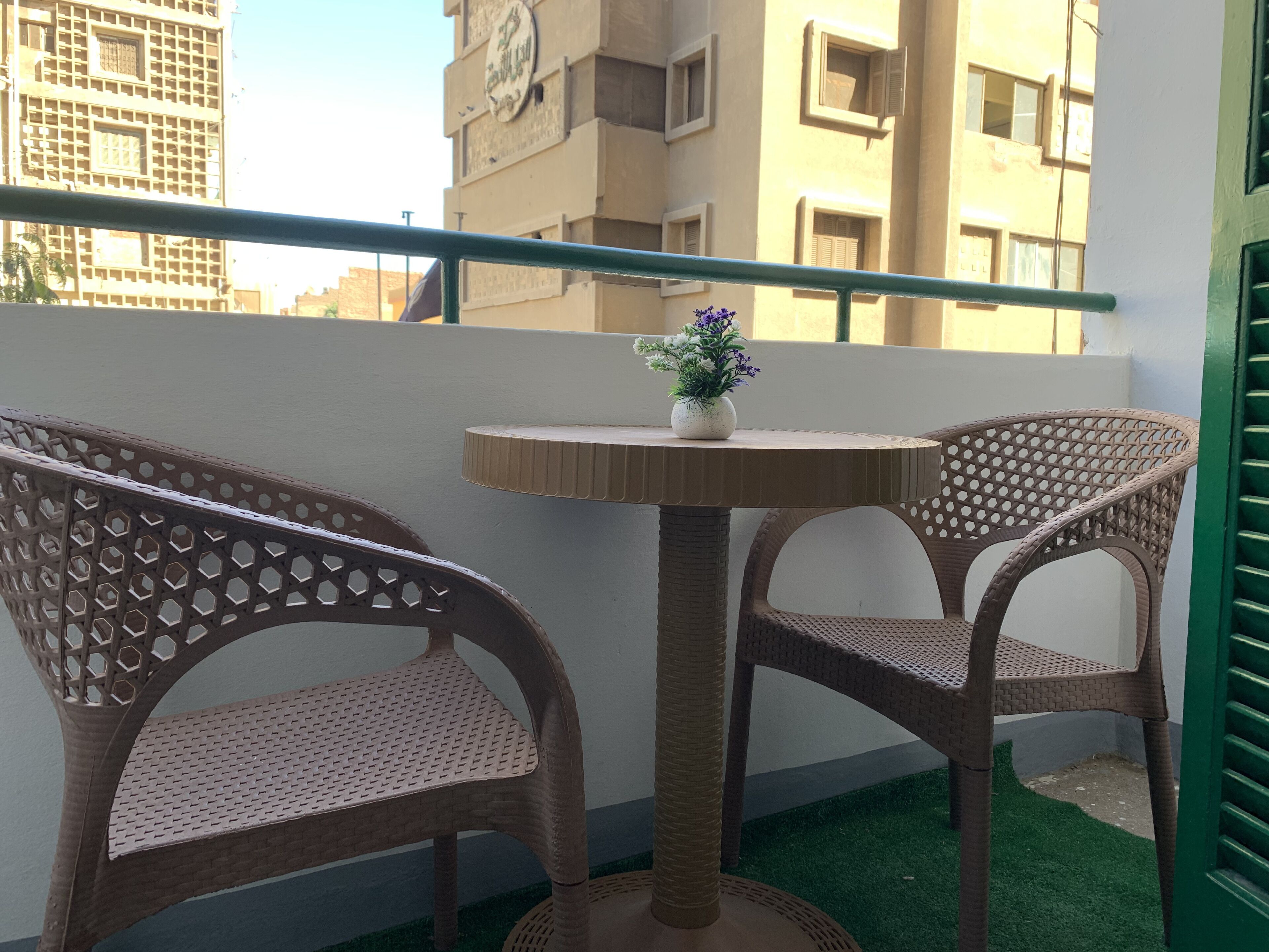 Un balcon avec deux fauteuils en osier, une petite table ronde ornée d’un vase de fleurs, et une vue sur les bâtiments qui se dressent à l’arrière-plan.