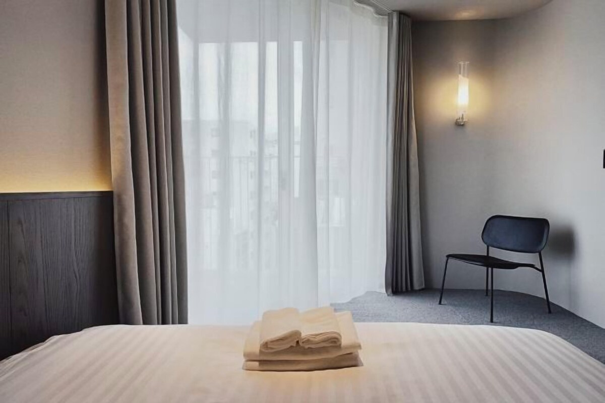A hotel room with a neatly made bed, a chair, and a wall-mounted light.