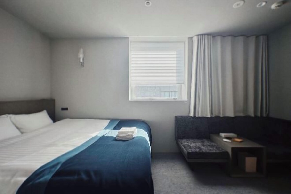 A hotel room with a bed, a small seating area, a window with blinds, and a nightstand.