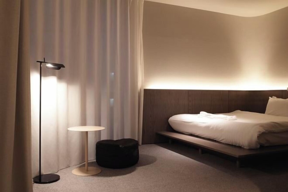 A modern hotel room with a bed, a round table, a floor lamp, and a black ottoman.