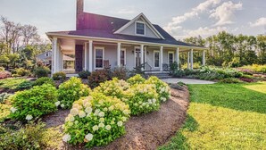A two-story house with a covered porch, surrounded by a well-maintained garden with various shrubs and flowers. - Mountain Blossom Retreat by Evergreen Home (Lanesborough)