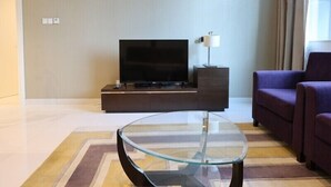 A modern living room with a flat-screen TV on a dark wooden stand, a glass coffee table, a purple sofa, and a lamp on a side table. - Cozy 1BR Buisness bay Damac Cour Jardin (Dubai)