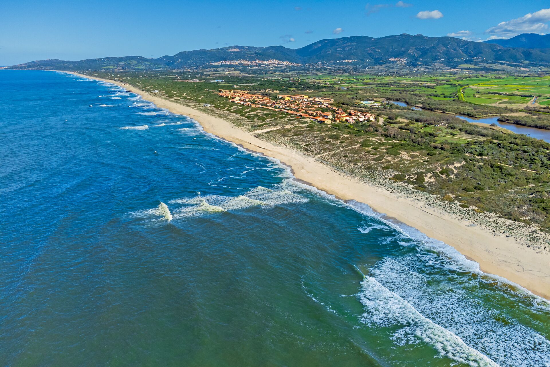 An aerial view of a coastal area with a beach, a small town, and mountains in the background.