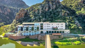 A white building with multiple arches and a wooden bridge leading to it, surrounded by lush greenery and mountains. - Apartment Bellavista Viddalba (Sardegna)