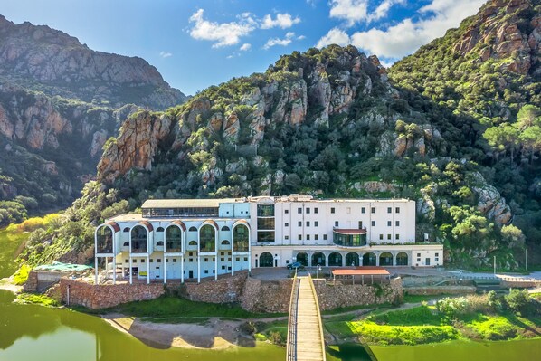 A white building with multiple arches and a wooden bridge leading to it, surrounded by lush greenery and mountains.