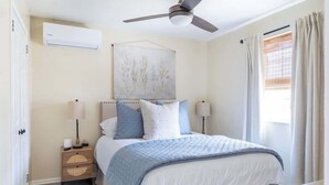 A bedroom with a bed, bedside lamps, a ceiling fan, an air conditioning unit, and a window with curtains.