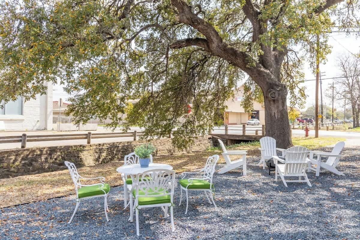 Un área de estar al aire libre con sillas blancas y una mesa, rodeada por un árbol y un camino de grava.
