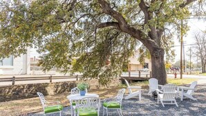 An outdoor seating area with white chairs and a table, surrounded by a tree and a gravel path.