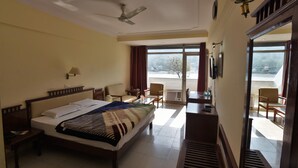 A hotel room with a bed, a desk, chairs, a ceiling fan, and a view of the outdoors. - Ganga Resort (Narendranagar)