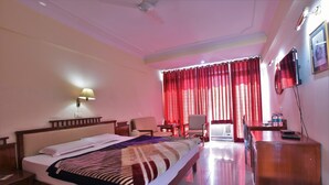 A hotel room with a bed, a desk, a chair, a television, and a window with red curtains. - Ganga Resort (Narendranagar)
