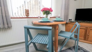 Une pièce avec une table en bois dressée pour deux personnes, une paire de chaises bleues, un vase avec des fleurs roses, une horloge au mur et un téléviseur posé sur une commode en bois.