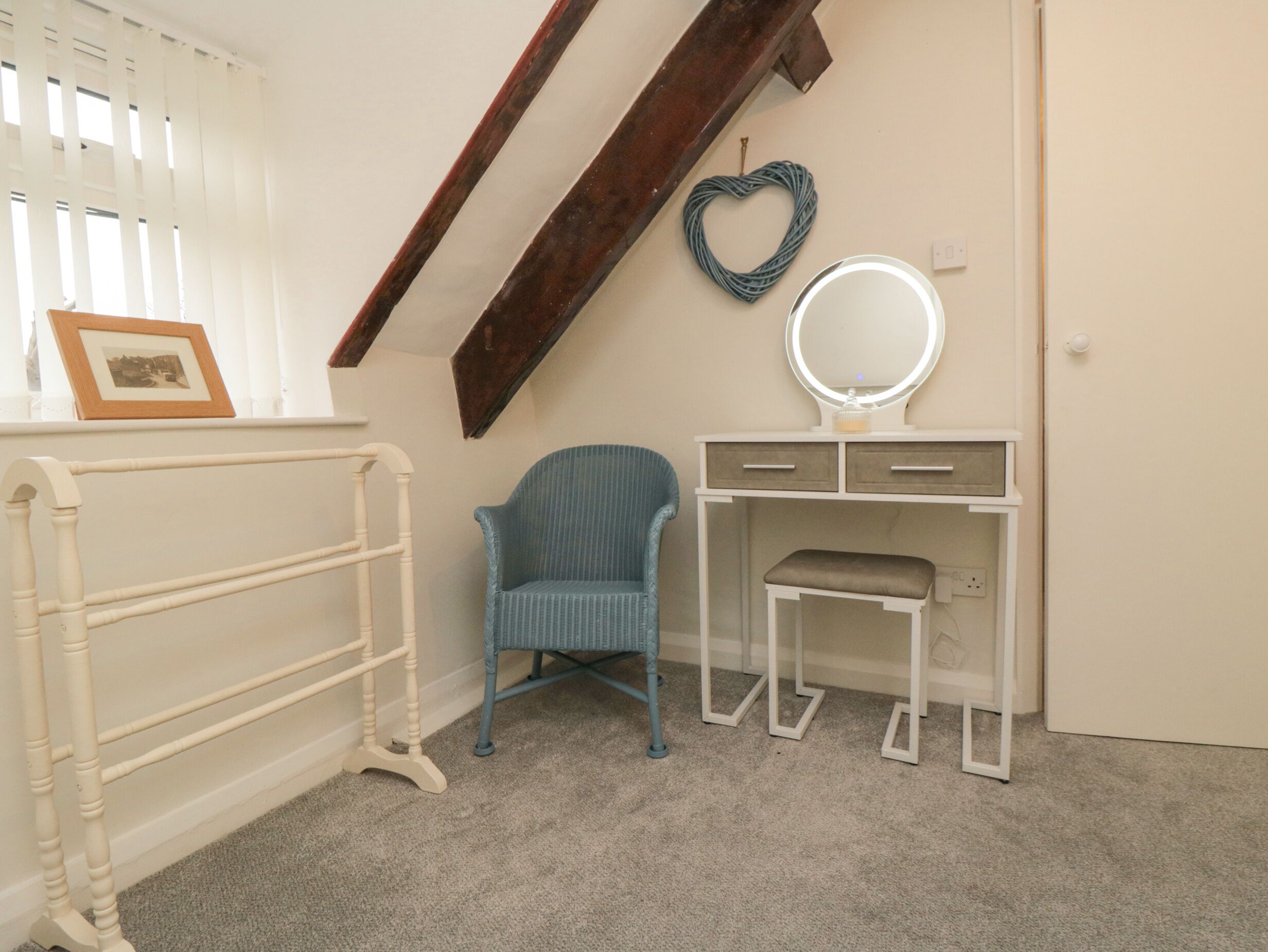 A room with a white vanity, a blue chair, a white bench, a wooden beam, a heart-shaped decoration, and a framed picture on the wall.