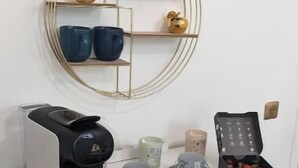 Una cafetera blanca sobre un mueble blanco, con un espejo de marco dorado encima.