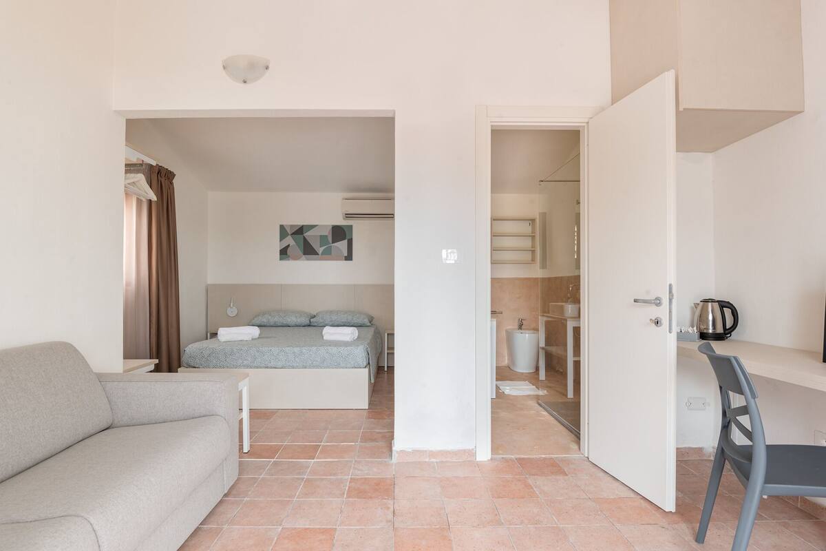 Une chambre d’hôtel moderne, équipée d’un lit, d’un canapé, d’un bureau et d’une salle de bain visible à travers une porte ouverte.