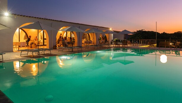 A pool area with lounge chairs and umbrellas, surrounded by covered seating areas with people dining. - Q Resort (Sorso)