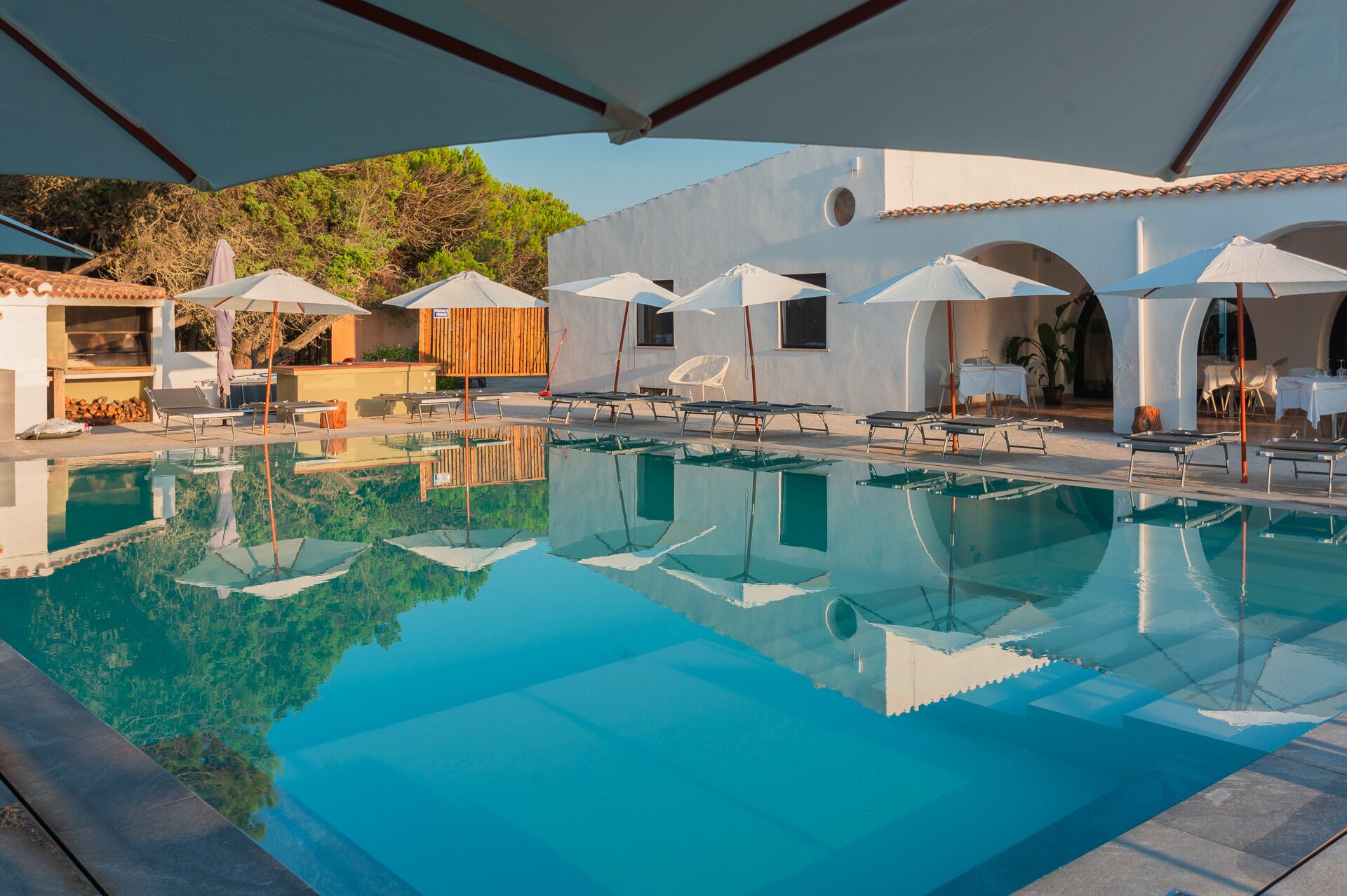 A clear swimming pool with lounge chairs and umbrellas, surrounded by white buildings with arches.