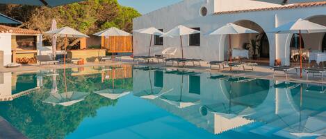 A clear swimming pool with lounge chairs and umbrellas, surrounded by white buildings with arches.