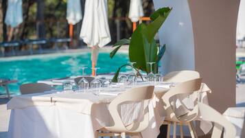 A dining table set for a meal with chairs, a pool in the background, and white umbrellas.