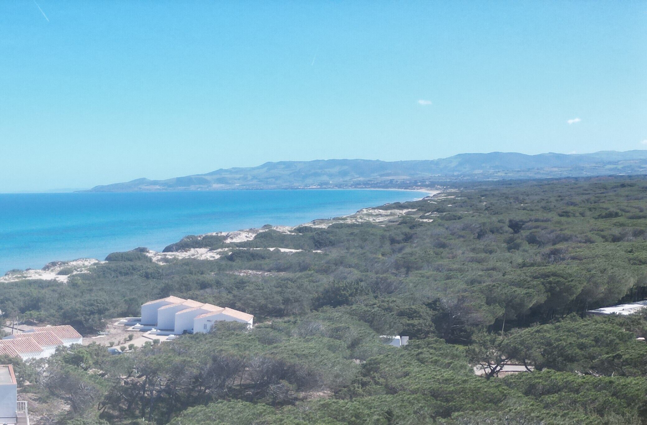 Un paisaje costero con una playa, abundante vegetación y un edificio con estructuras cilíndricas grandes.