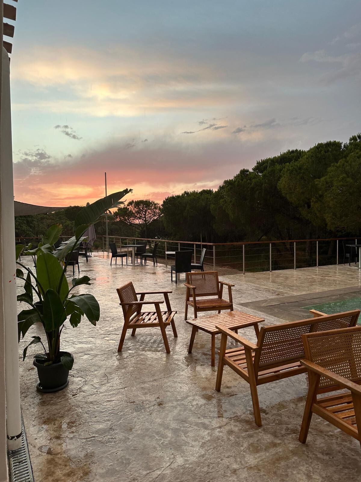 An outdoor seating area with wooden furniture and a view of the sunset over a forested area.