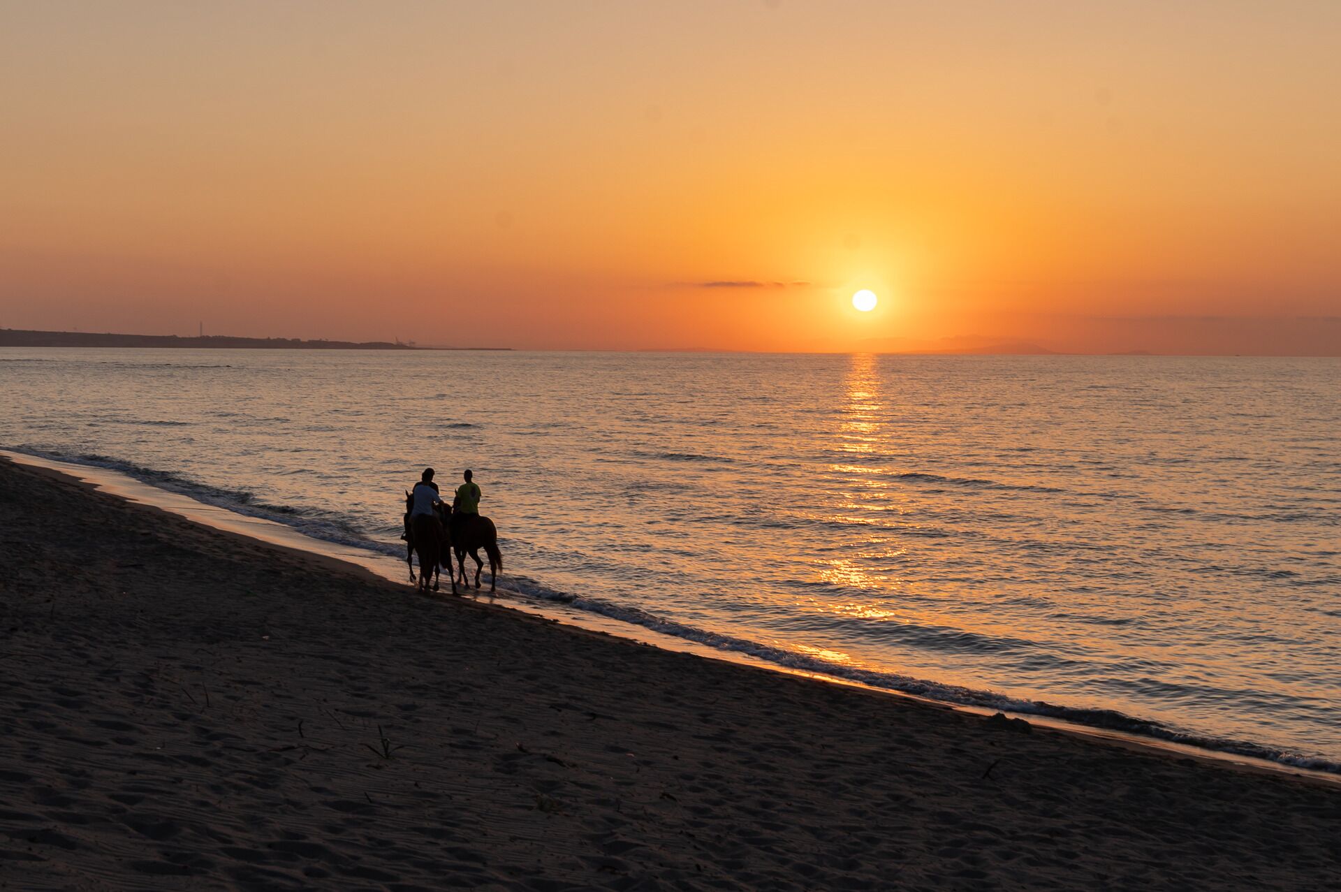 A sunset view with two people on horseback on a beach.