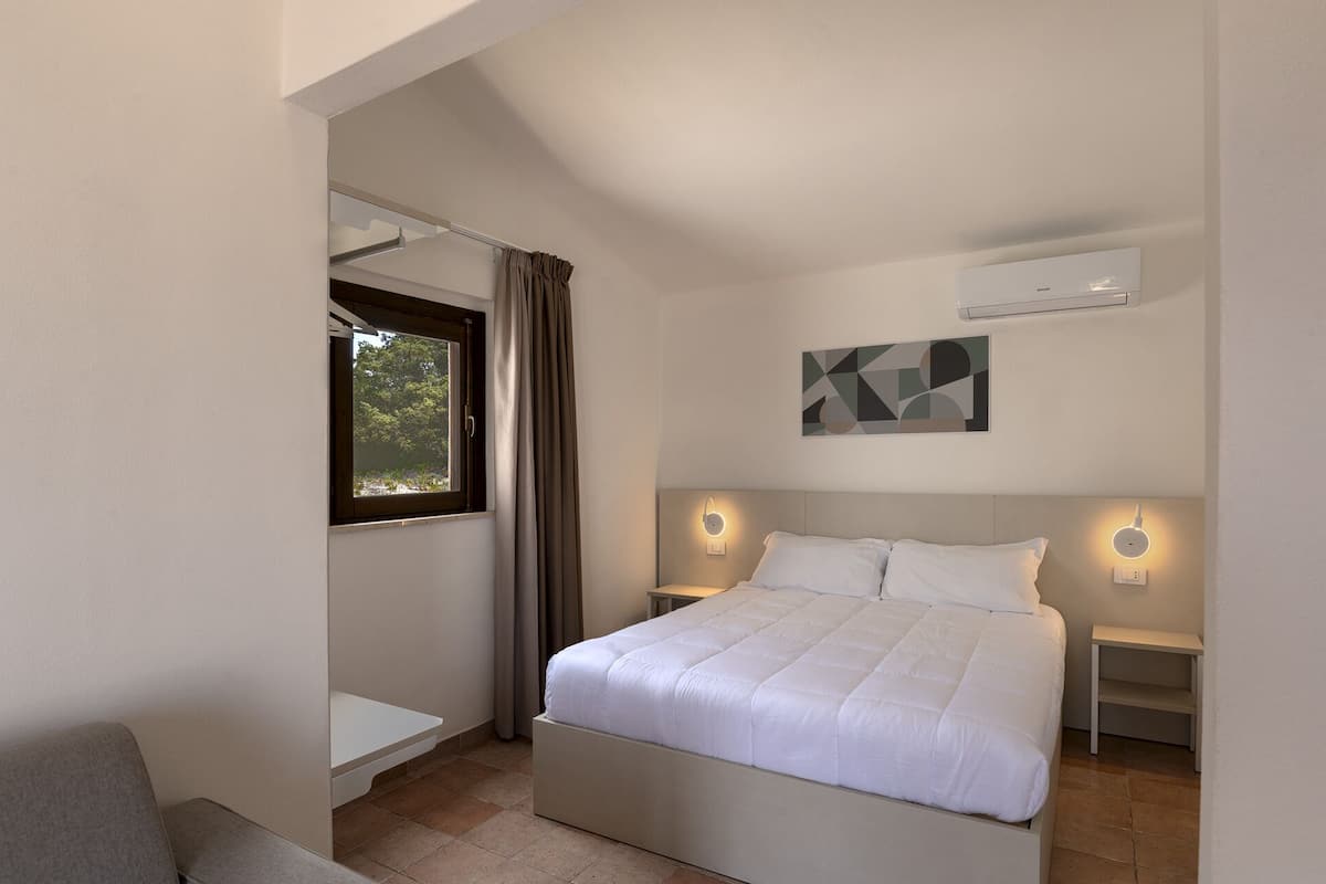 Une chambre moderne avec un lit, des tables de chevet, une fenêtre avec des rideaux, un climatiseur fixé au mur et une œuvre d’art aux motifs géométriques.