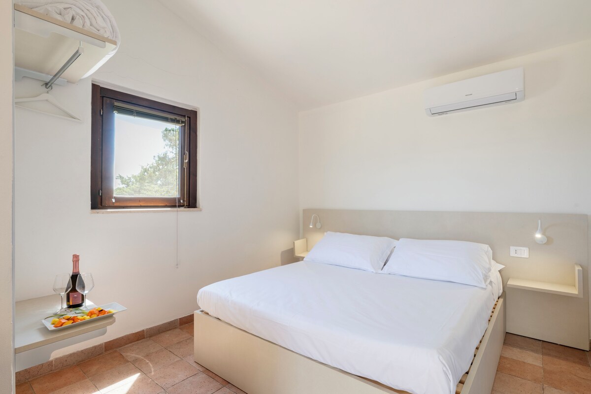 A bedroom with a bed, a bedside table with a bottle and plate, a window, and an air conditioning unit.
