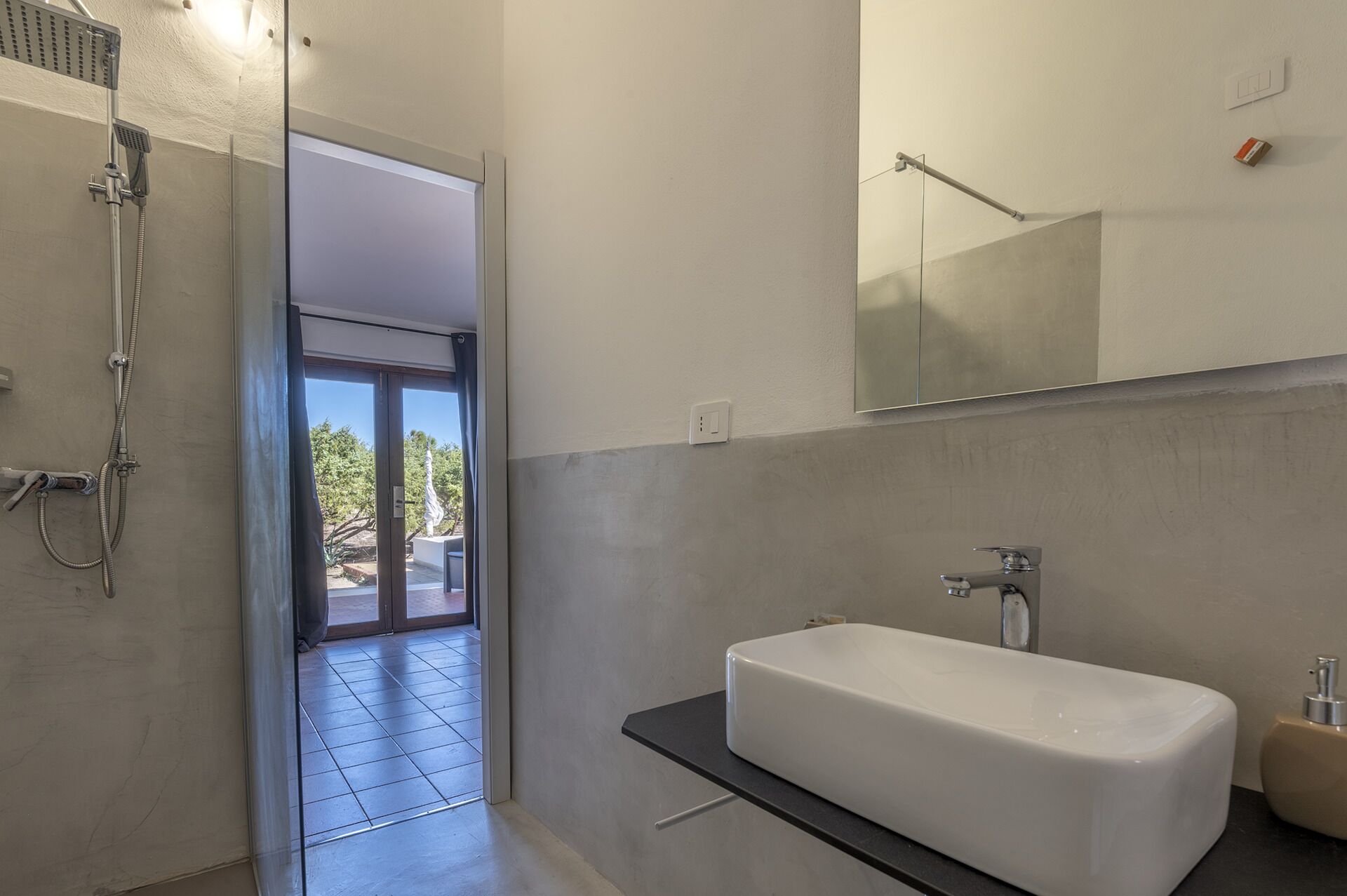 Un bagno moderno con una vasca da bagno bianca ampia, un box doccia in vetro e vista su un giardino attraverso la porta in vetro.