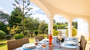 Un espace de restauration en plein air avec une table dressée pour quatre personnes, entourée de chaises en osier, sous une structure couverte soutenue par des colonnes.
