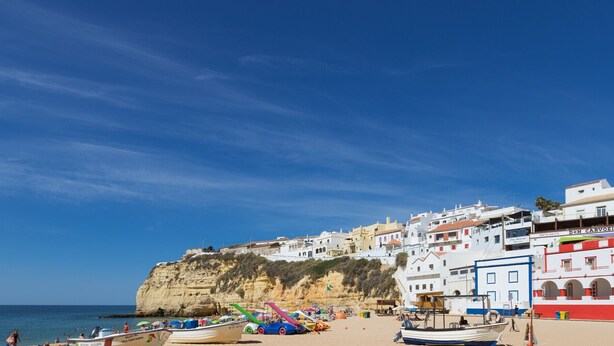 Une ville côtière avec des bâtiments construits le long de la falaise, une plage parsemée de parasols et de bateaux, et un ciel bleu dégagé.