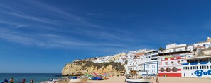 Une ville côtière avec des bâtiments construits le long de la falaise, une plage parsemée de parasols et de bateaux, et un ciel bleu dégagé.