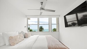 A bedroom with a bed, a ceiling fan, a TV mounted on the wall, and a large window with a view of the ocean. - Azure Breezes | Modern Sanctuary by the Beach (Carolina)