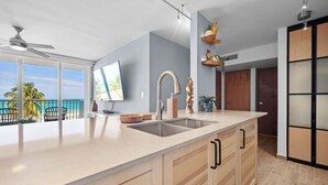 A modern kitchen with a large island, a built-in oven, and a view of the ocean. - Azure Breezes | Modern Sanctuary by the Beach (Carolina)