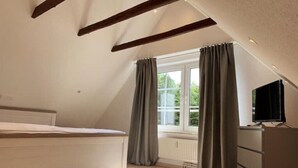 A bedroom with a sloped ceiling, a bed, a dresser, a television, and a window with curtains. - Ferienhaus Muschel mit Garten an der Nordsee (Dornum)