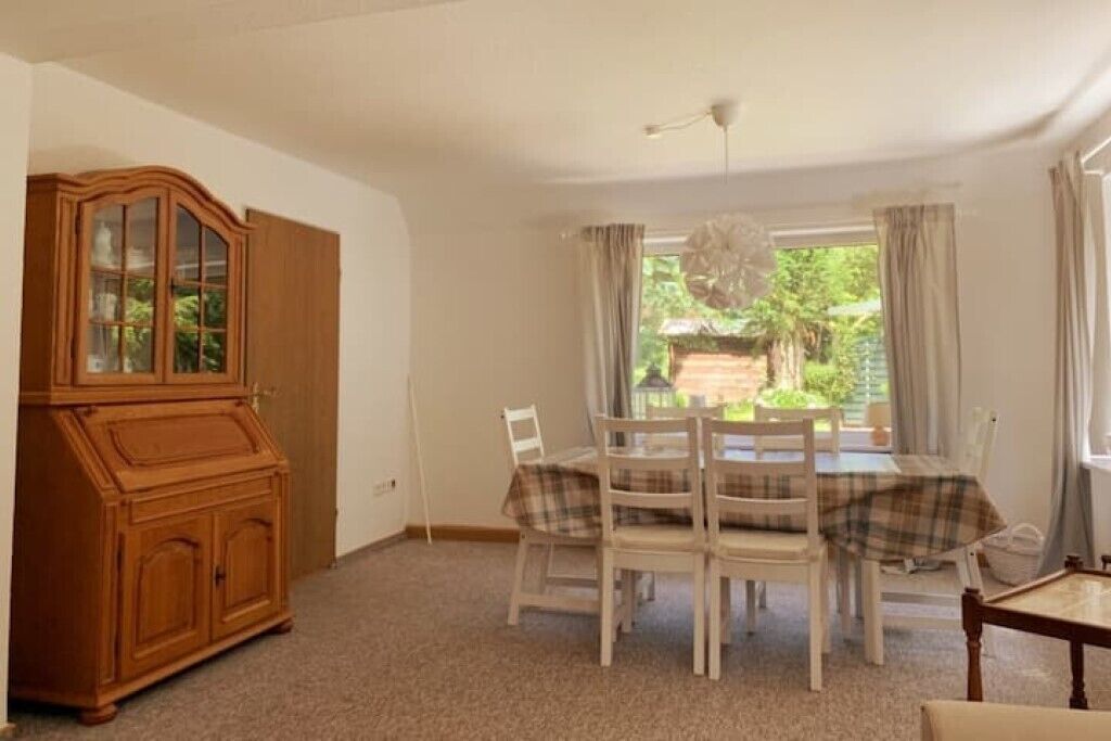 A dining area with a table set for four, surrounded by white chairs, a wooden cabinet, and a view of greenery outside.