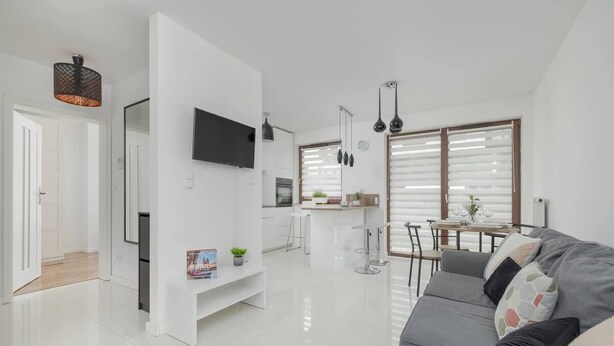A modern living room with a grey sofa, a flat-screen TV mounted on the wall, and a dining area with a table and chairs.