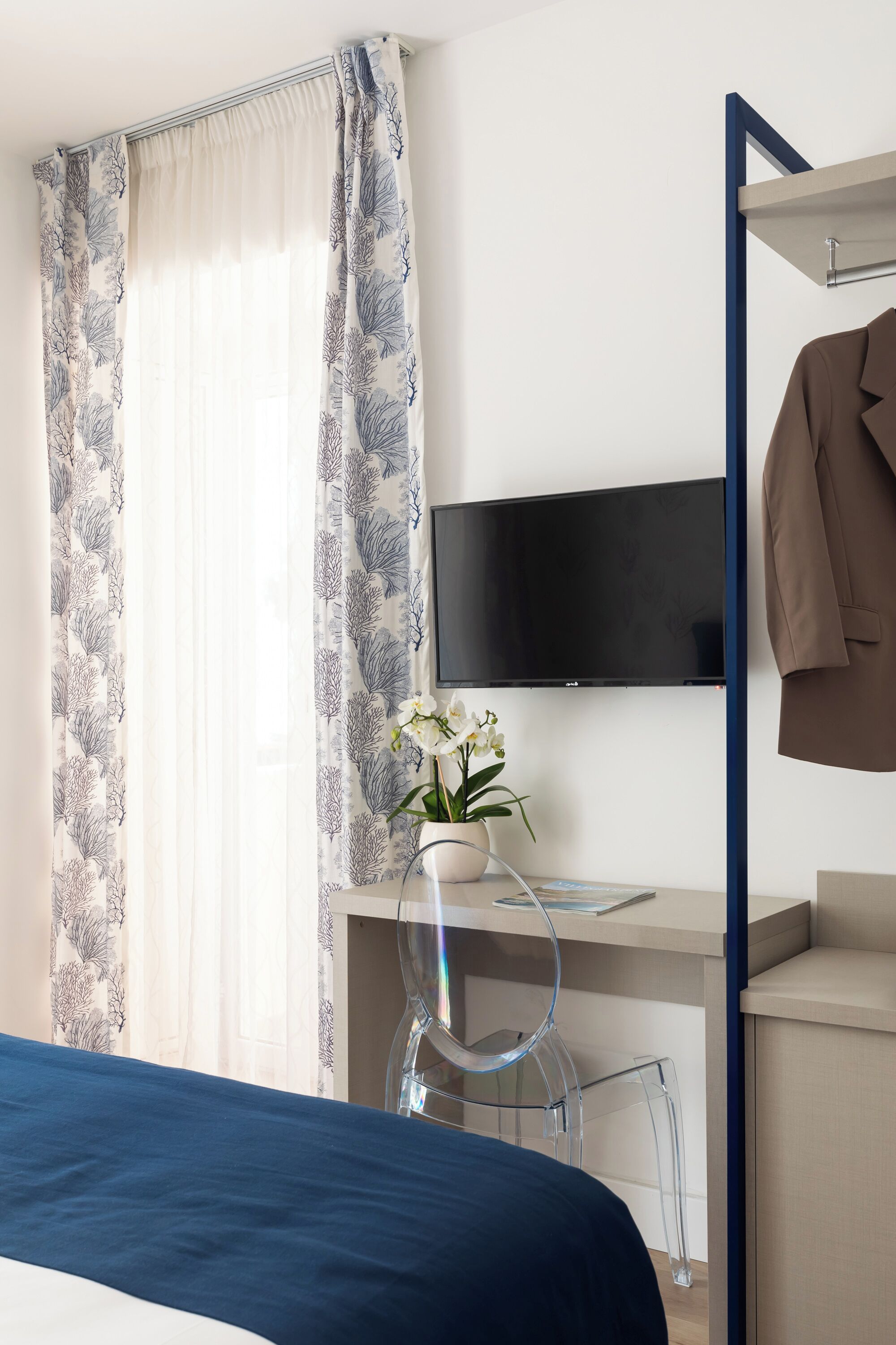 Quarto de hotel com uma cama, uma escrivaninha com cadeira, televisão na parede e um cabideiro com um casaco.
