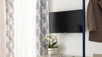 Quarto de hotel com uma cama, uma escrivaninha com cadeira, televisão na parede e um cabideiro com um casaco.