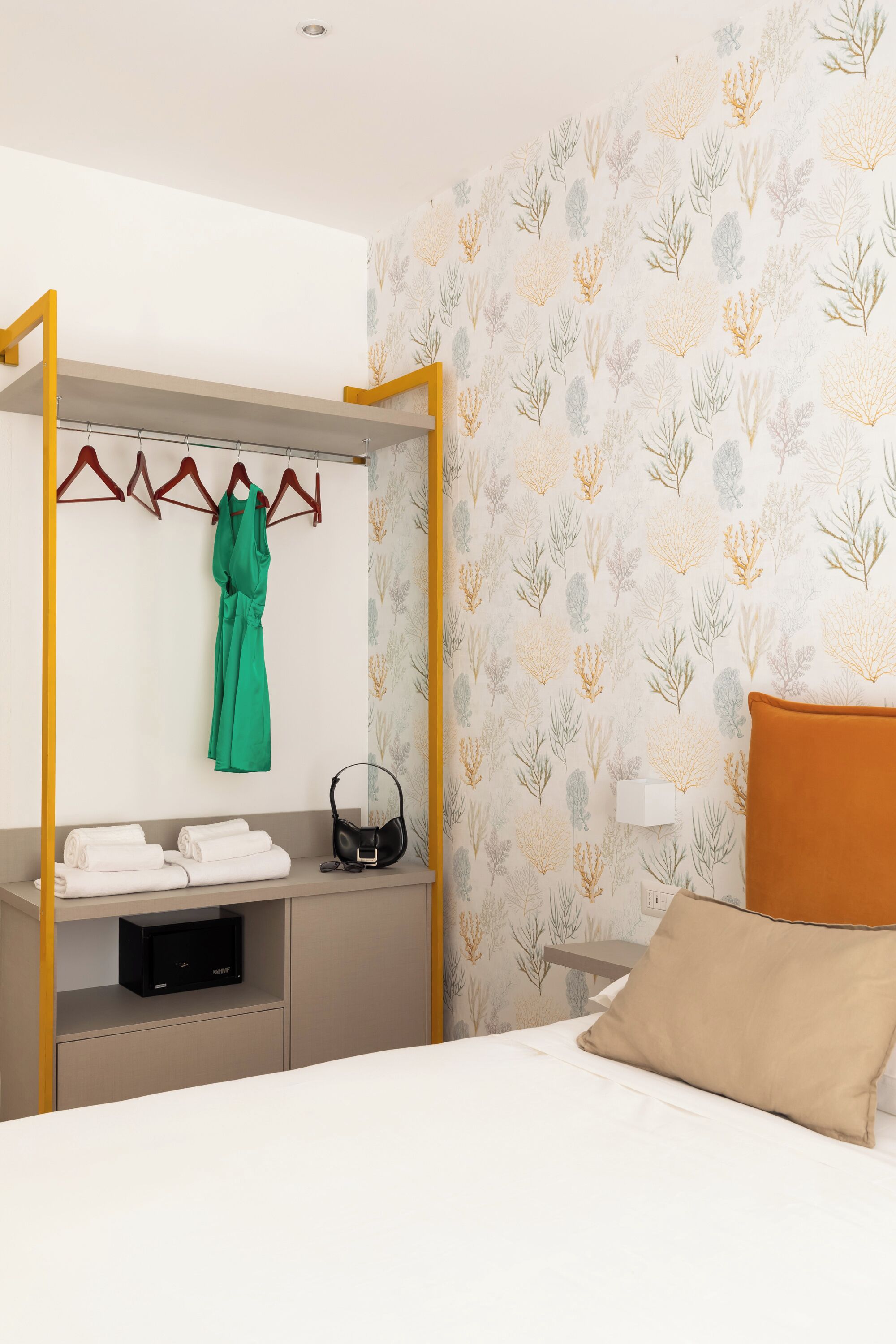 A hotel room with a bed, a wardrobe with a hanging robe, a bedside table with a lamp, and a wall with a leaf pattern.