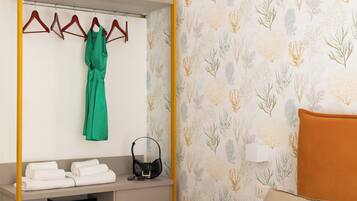 A hotel room with a bed, a wardrobe with a hanging robe, a bedside table with a lamp, and a wall with a leaf pattern.