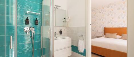 Un baño moderno con cabina de ducha de vidrio, un lavabo blanco con espejo, y una cama con cabecera naranja en la habitación contigua.