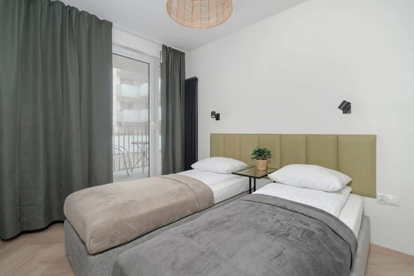 A cosy space with white walls and long curtains. Two single beds with olive-coloured headboard.