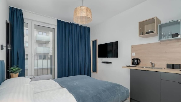 A modern bedroom with a bed, a TV, a kitchen area, and a balcony with blue curtains.