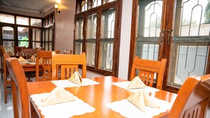 Un espace repas avec des tables et des chaises en bois, de grandes fenêtres équipées de moustiquaires et une vue sur l’extérieur.