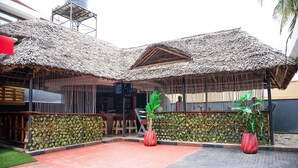 Un espace extérieur abrité, doté d’un toit de chaume, d’une clôture en bambou et de deux grands pots rouges contenant des plantes.