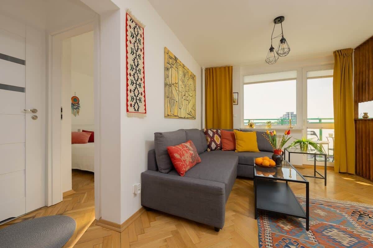 A modern living room with a grey sofa, a coffee table, and a patterned rug. There are two windows with curtains, a door, and a wall with a decorative tapestry.