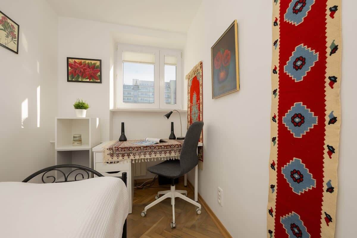 Une chambre à coucher avec un lit, un bureau avec un ordinateur, une chaise, une fenêtre et un rideau rouge à motifs.