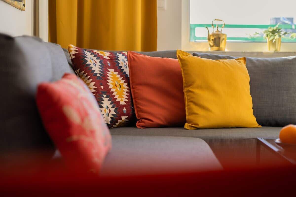 A modern living room with a grey sofa, colorful cushions, and a view of a cityscape through a window.