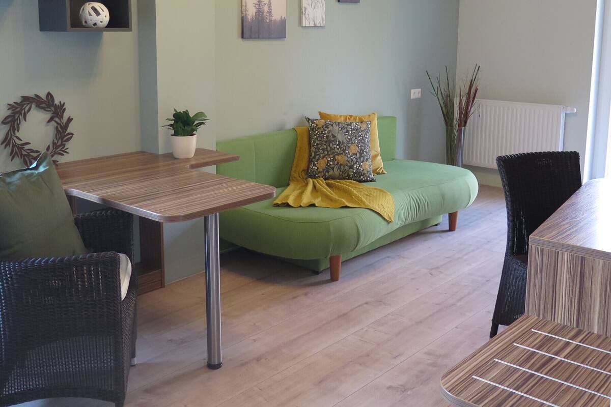 A modern living room with a green sofa, a wooden table, and a green chair. There are wall-mounted shelves with decorative items and a radiator.