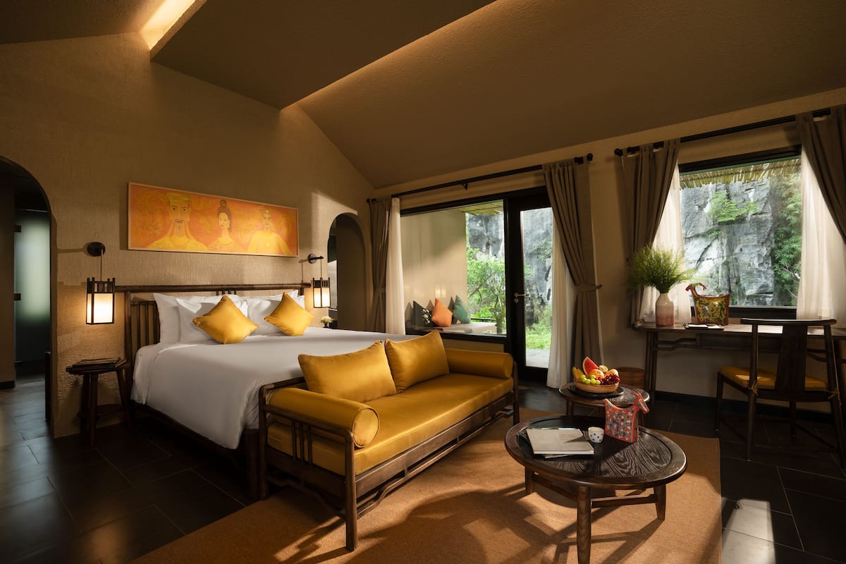 Habitación de hotel con cama, sofá, mesa de comedor y vista a zonas verdes por la ventana.